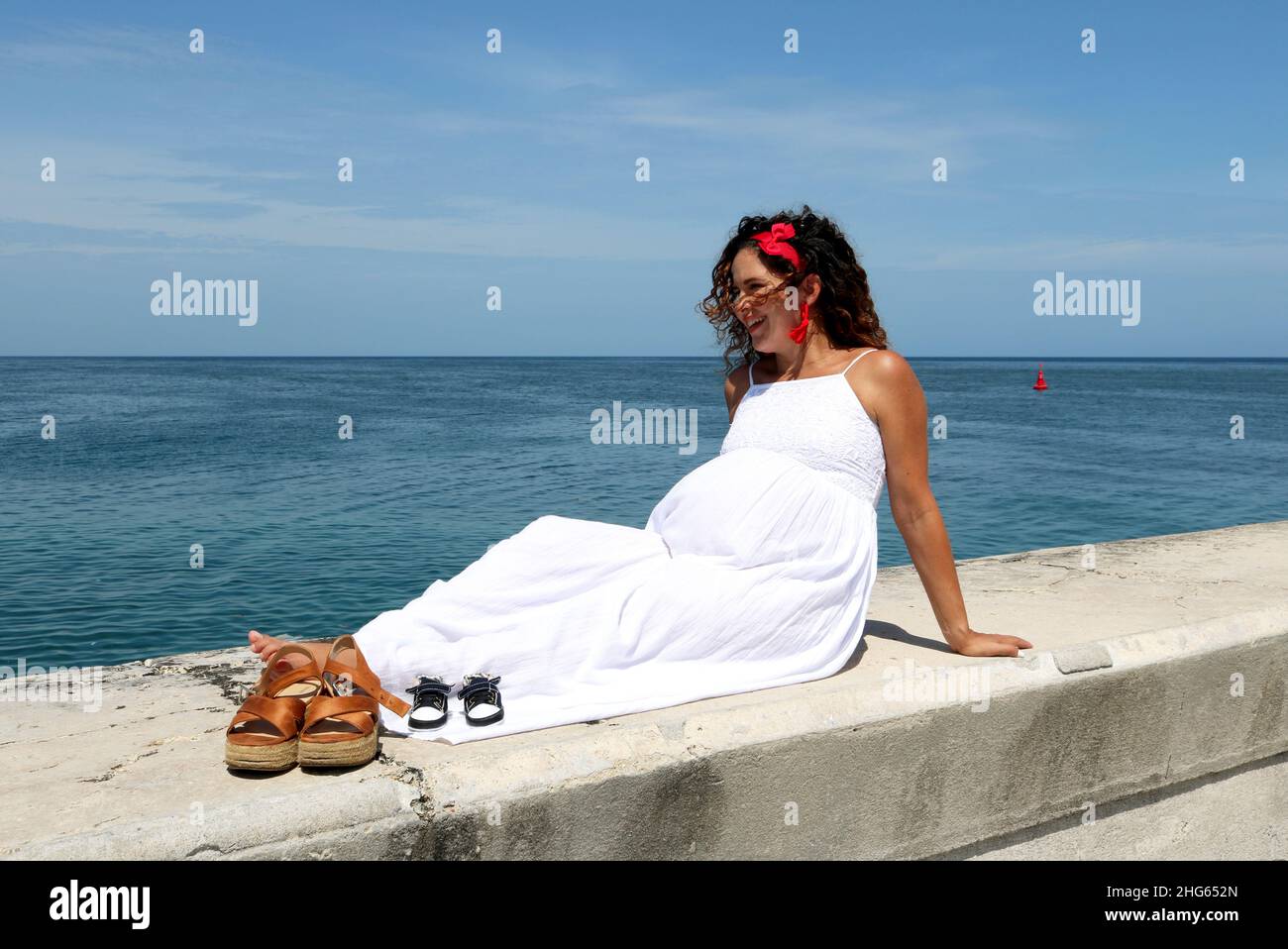 Donna incinta seduta rilassata vicino all'oceano godendo l'aria fresca, il sole e la vista dell'oceano. Le sue scarpe accanto al suo bambino atteso Foto Stock