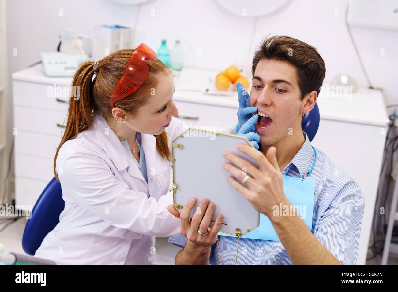 Da sopra donna rossa in accappatoio bianco che tocca i denti dell'uomo con specchio durante l'appuntamento in clinica dentale moderna Foto Stock
