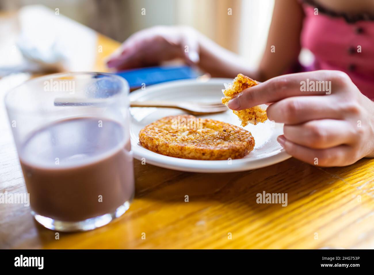 Macro closeup di donna seduta al tavolo della colazione che tiene mangiare patate fritte hash browns con mano su piatto da bicchiere di latte al cioccolato e sfondo Foto Stock