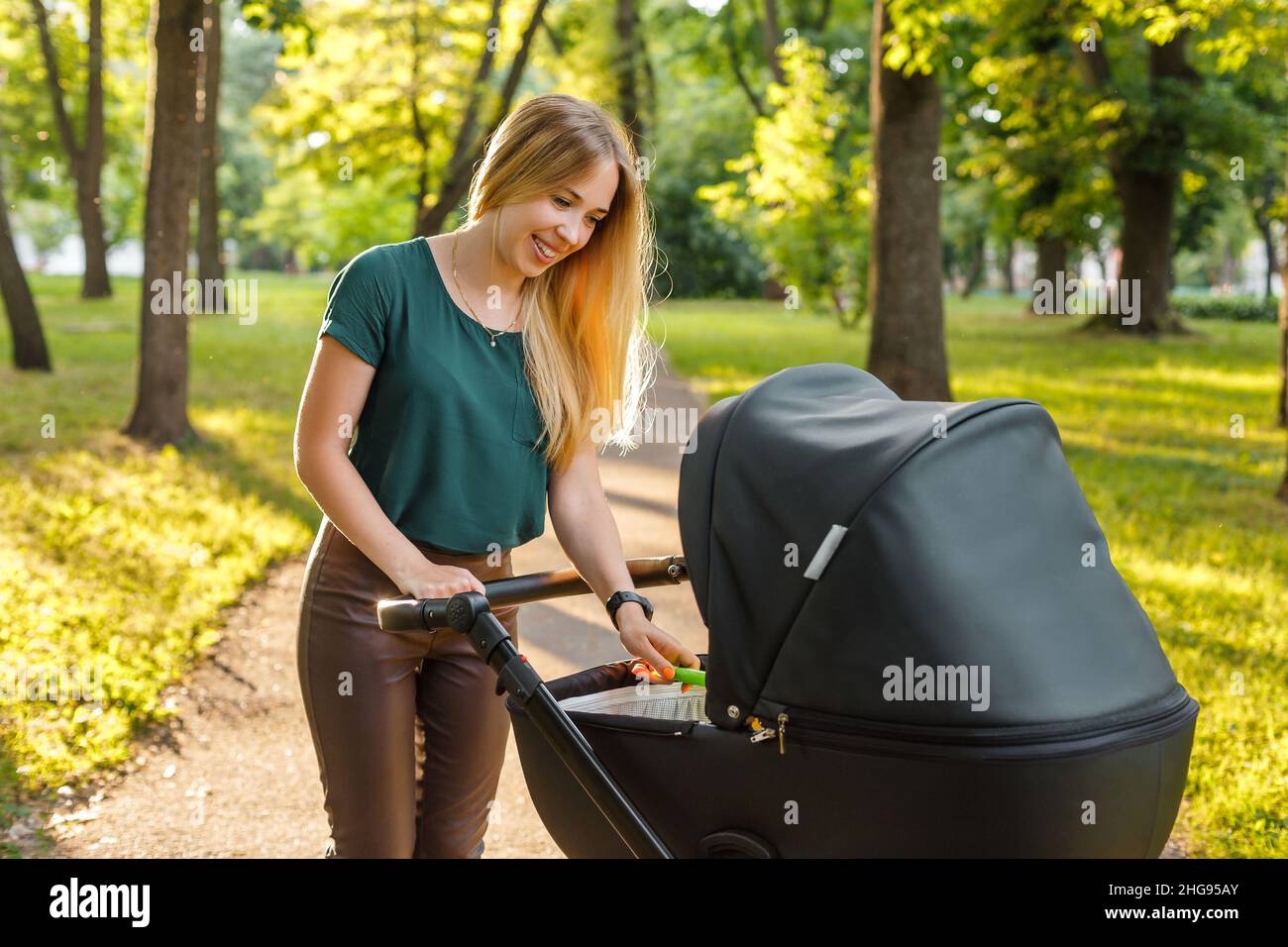 Giovane donna bionda che cammina con passeggino nero nel parco estivo. Mamma felice con il bambino in pam all'aperto. Foto Stock