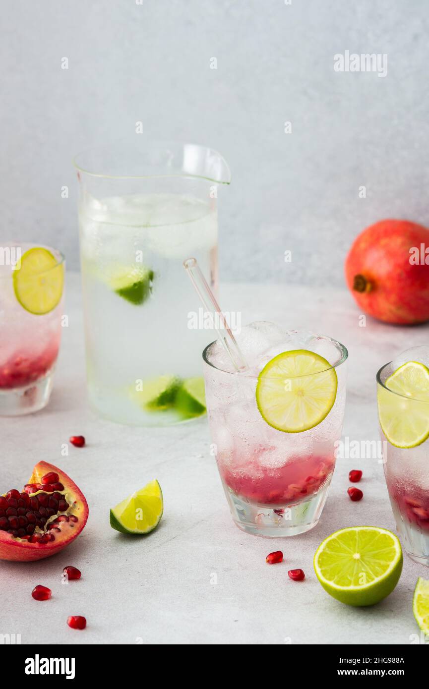 Rinfrescante bevanda di melograno in bicchieri con semi di melograno e ghiaccio, cannucce di calce e vetro, brocca di acqua fredda con agrumi, melograni tagliati Foto Stock