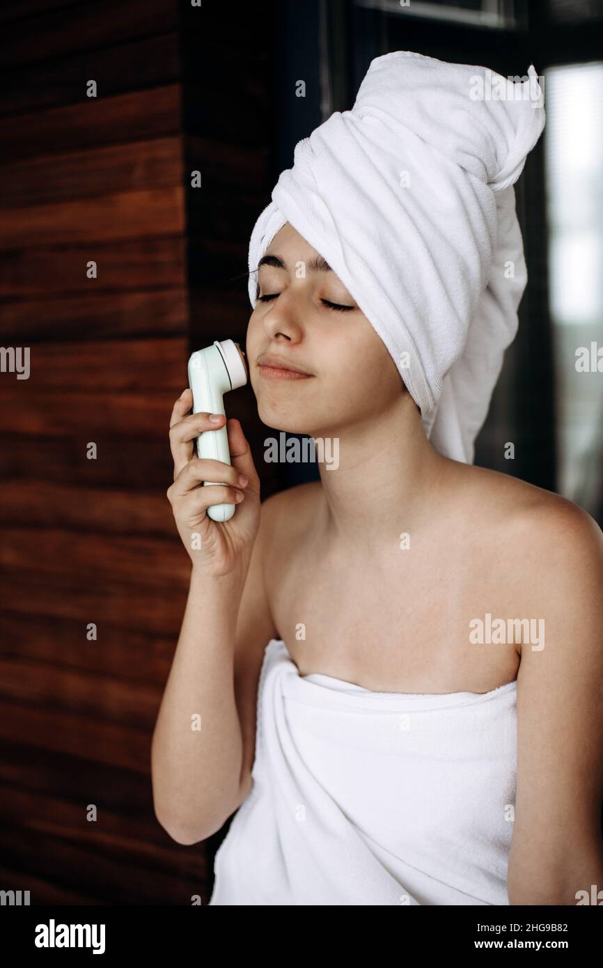 La bambina con asciugamano sulla testa fa il massaggio facciale con il massaggiatore elettrico senza fili Foto Stock
