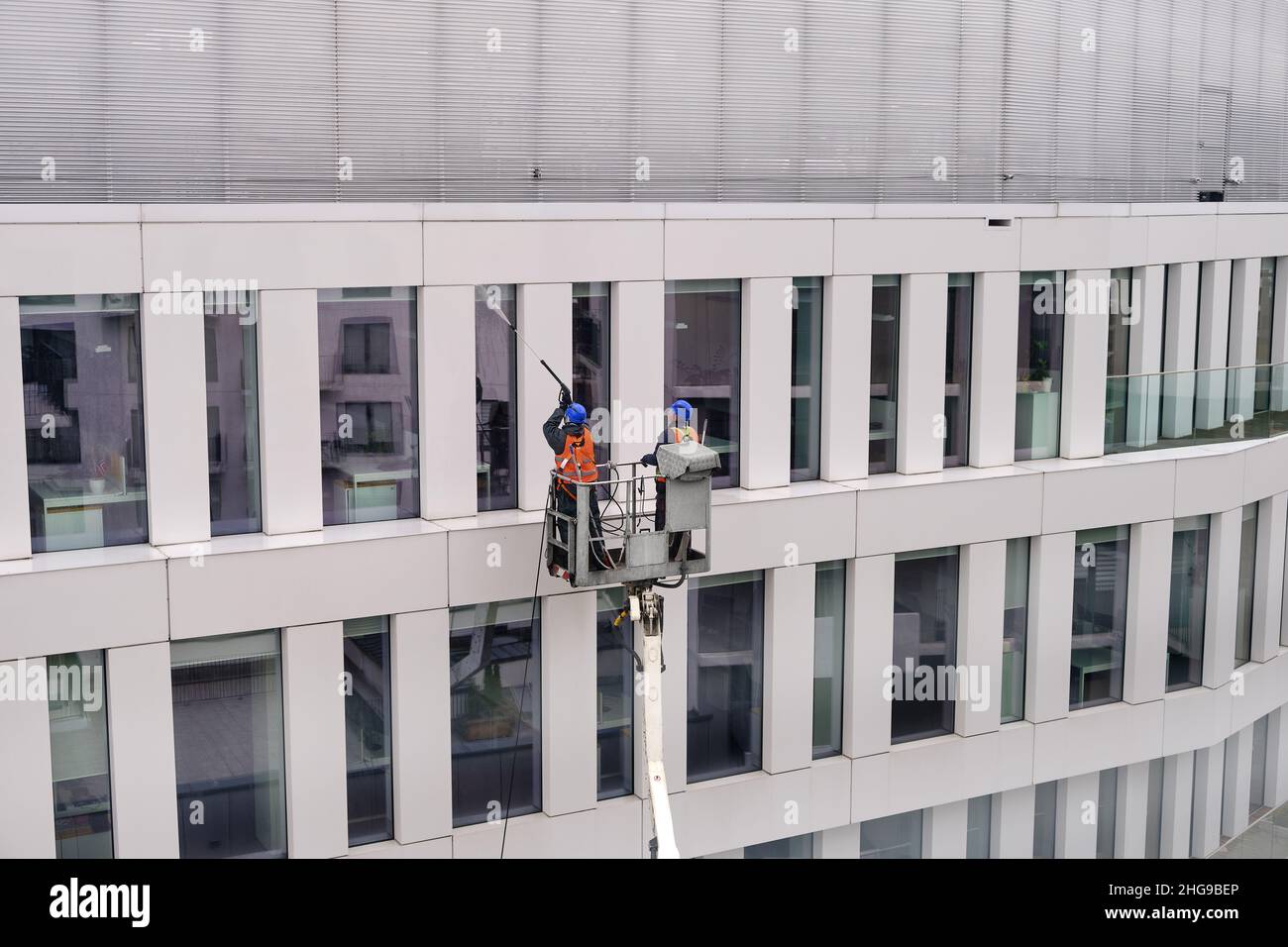 Due lavoratori che indossano imbracature di sicurezza lavano la facciata dell'edificio in altezza in piedi in una culla della gru o piattaforma aerea utilizzando idropulitrici a pressione e spazzatrici Foto Stock