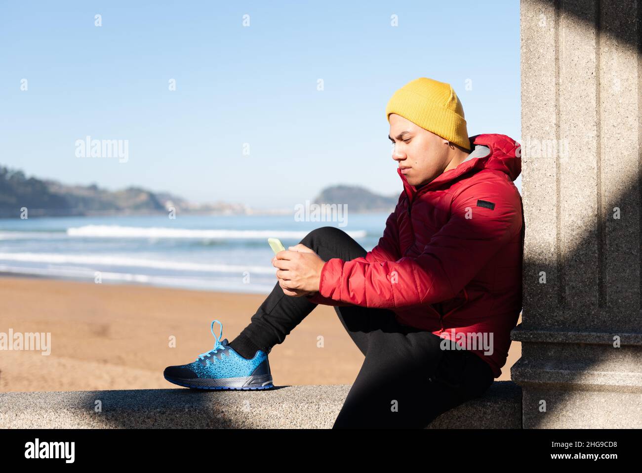 L'uomo latino si concentrò sul telefono mentre era seduto al mare. Concetto di dipendenza digitale. Foto Stock