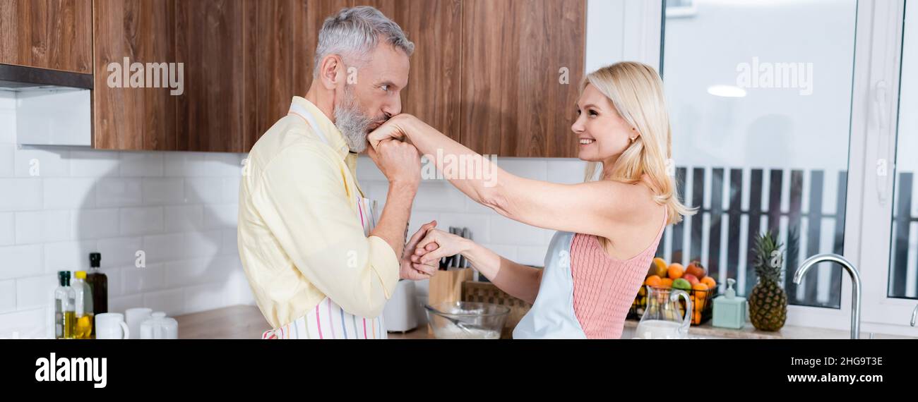 Uomo di mezza età in grembiule baciando la mano di sorridente moglie in cucina, banner Foto Stock