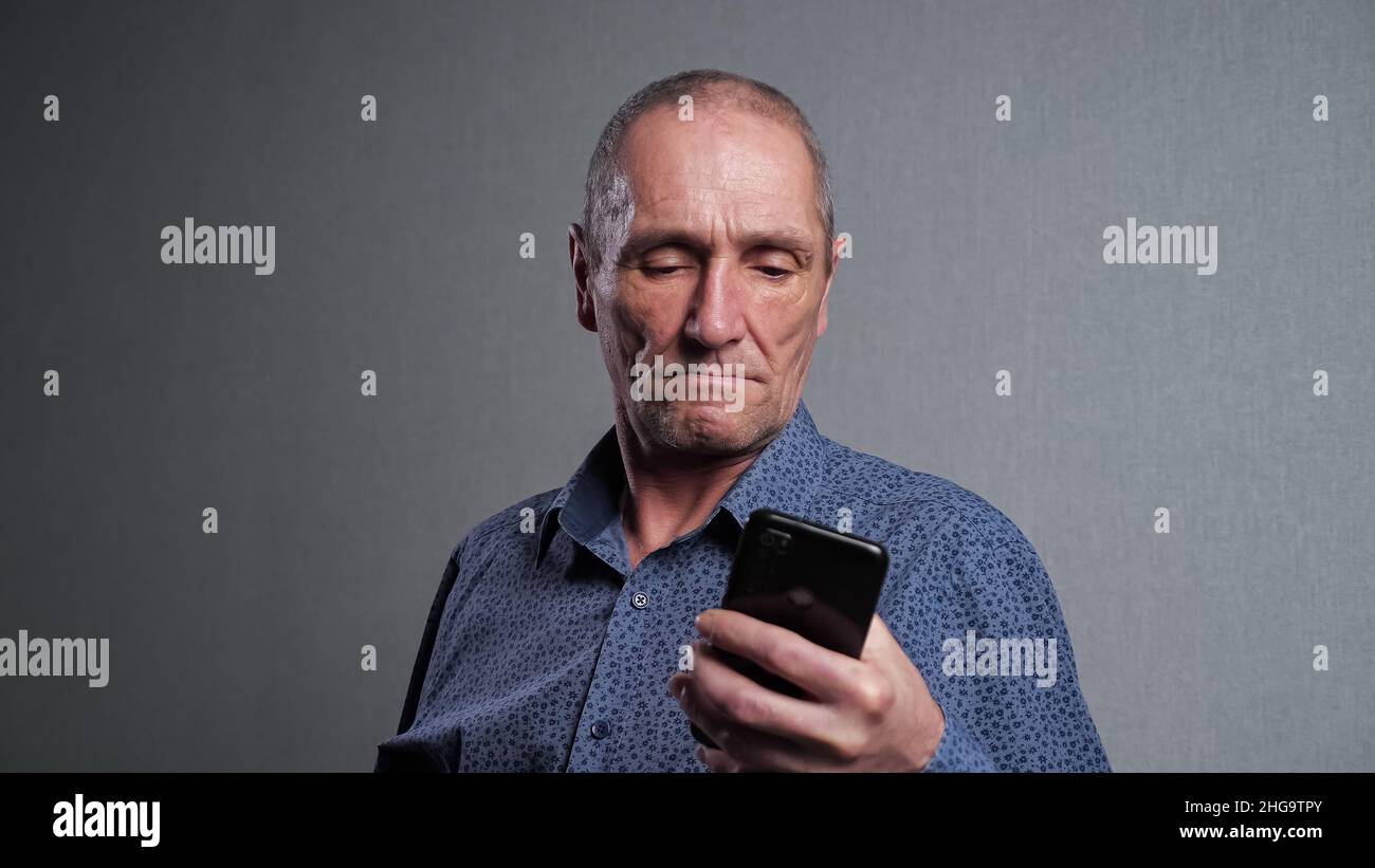 L'uomo anziano legge le notizie nel telefono mobile su sfondo grigio Foto Stock