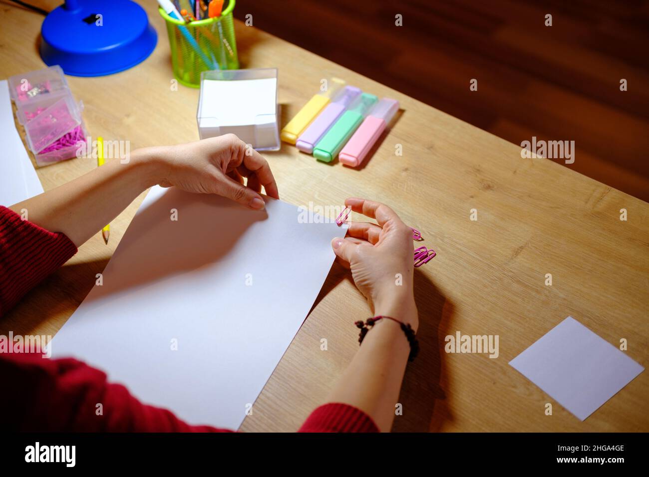 Donna che lavora nel suo ufficio su tavolo in legno a casa, vista dall'alto di un tavolo con attrezzature di studio e luce. Lady collega le graffette alle pagine Foto Stock