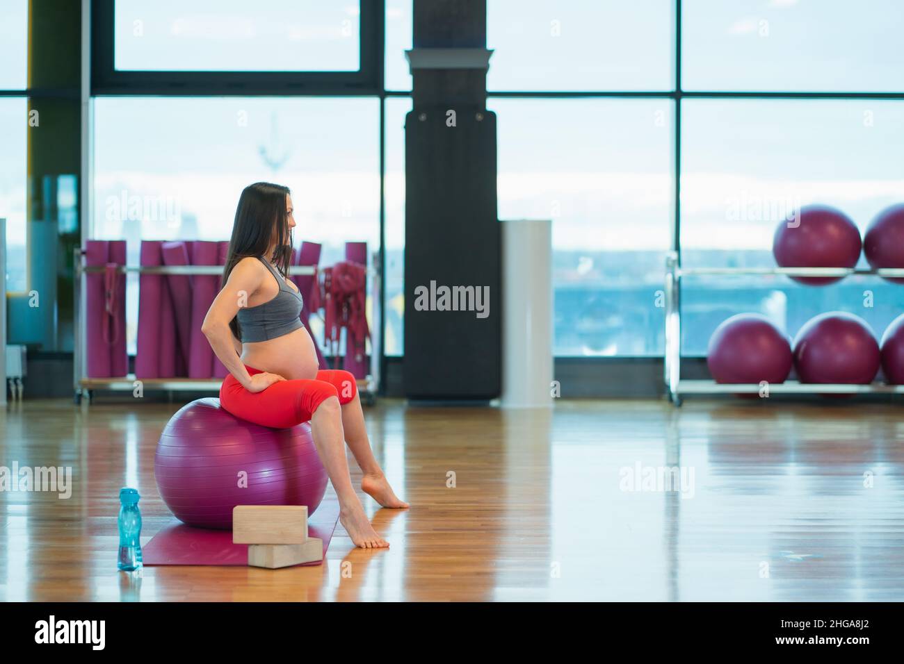 Vista laterale della giovane donna incinta in abbigliamento sportivo seduta su fit ball esercizio all'aperto in palestra. Foto Stock