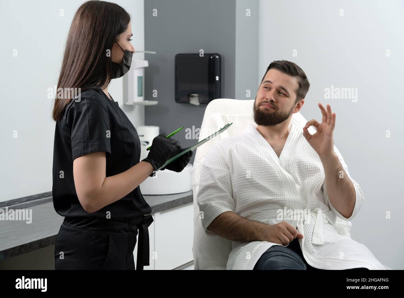 Cosmetologa femminile esperta che dà la consultazione al giovane paziente seduto su un divano in ambulatorio estetista di clinica estetica Foto Stock