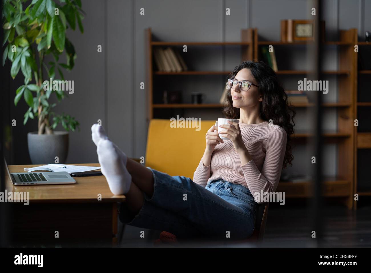 Giovane dipendente femminile rilassato che riposa sul posto di lavoro con una tazza di caffè e le gambe sul tavolo in ufficio Foto Stock