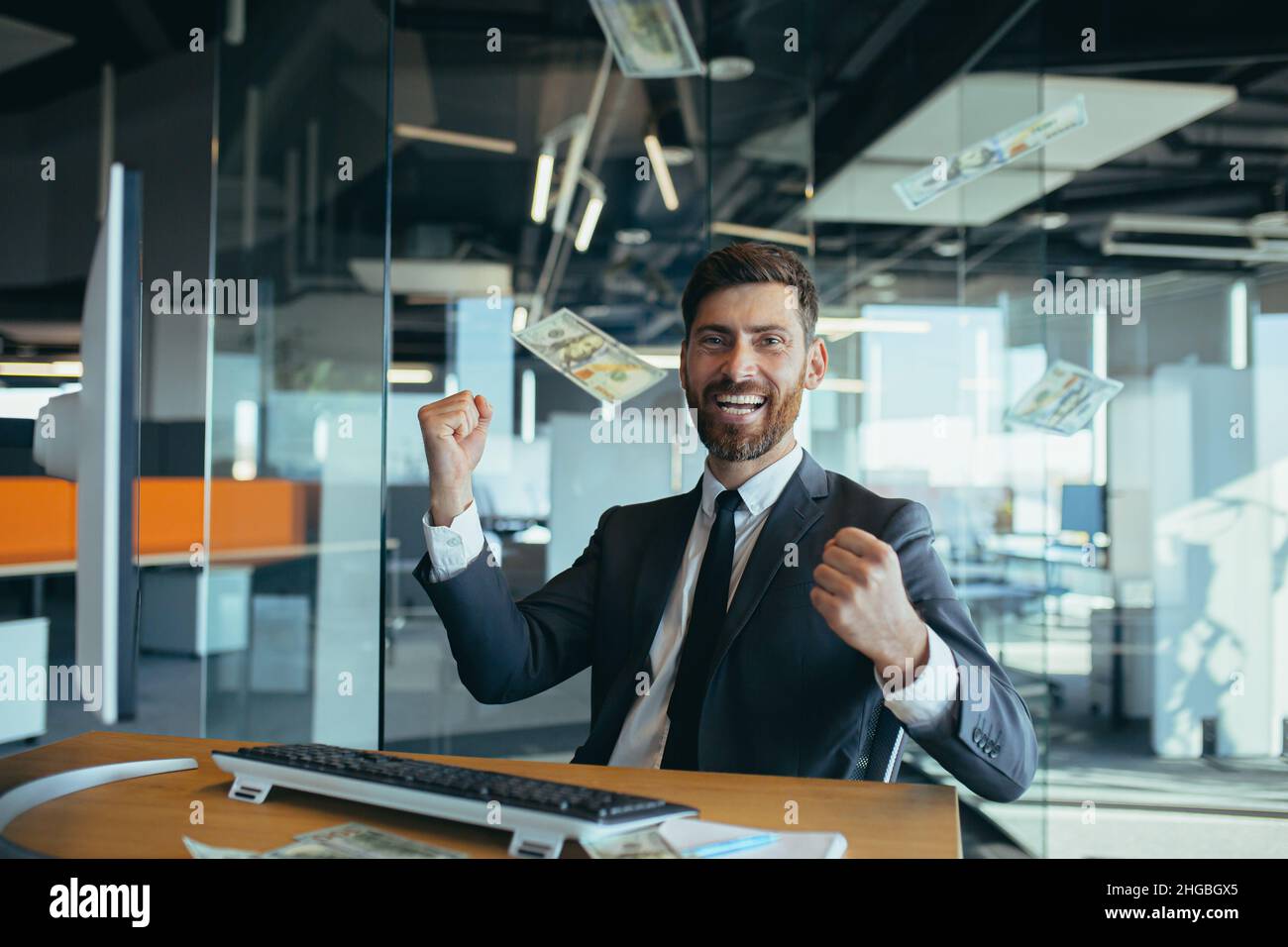Bel giovane uomo d'affari gioioso seduto su una scrivania, lavorando in un ufficio moderno, gettando soldi sulla testa. L'uomo sorridente felice getta soldi nella A. Foto Stock