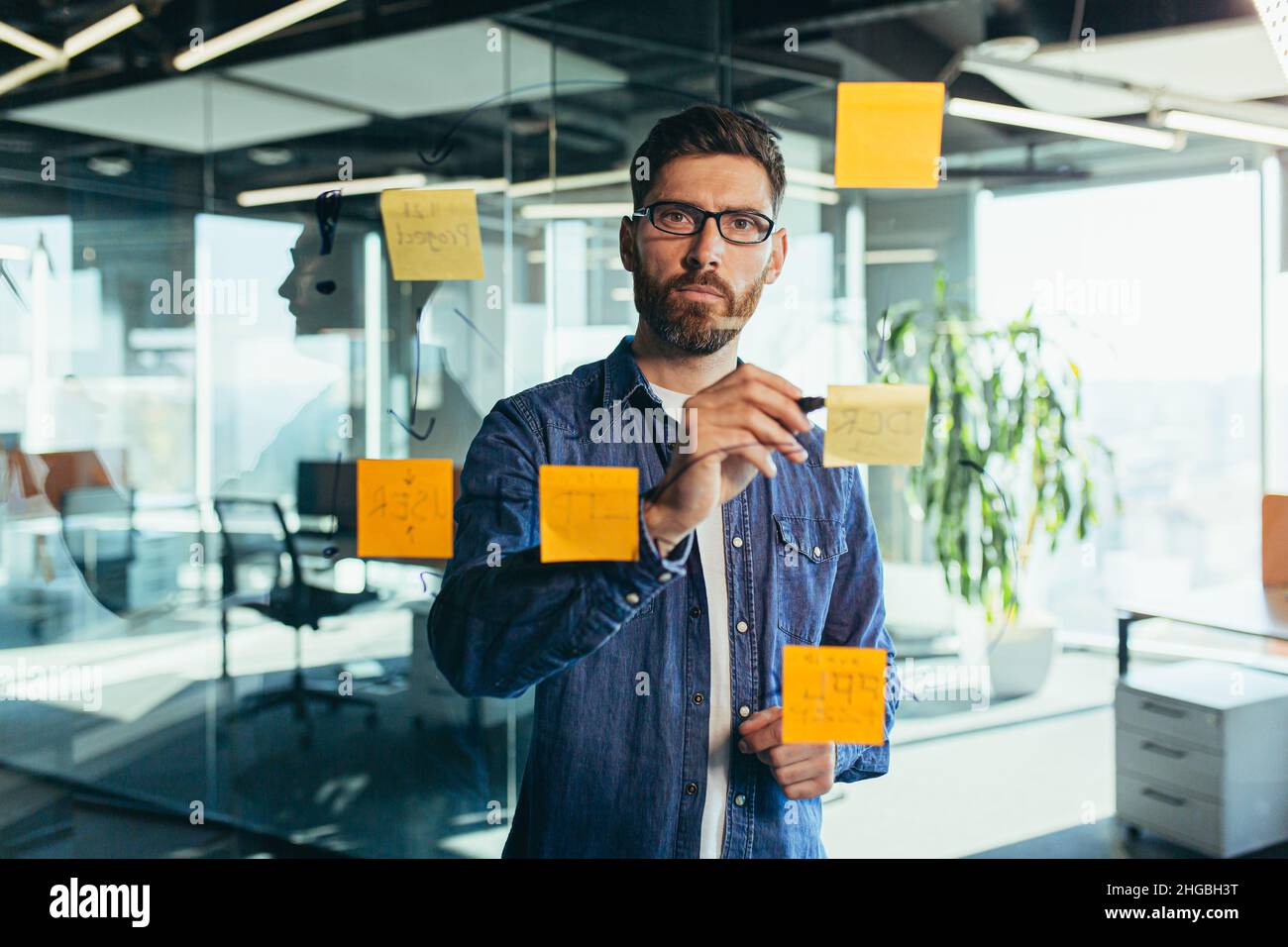 Uomo d'affari senior focalizzato in abiti casual scrittura su adesivo con marcatore nero. Note colorate appiccicate su una parete di vetro in una sala conferenze. Foto Stock