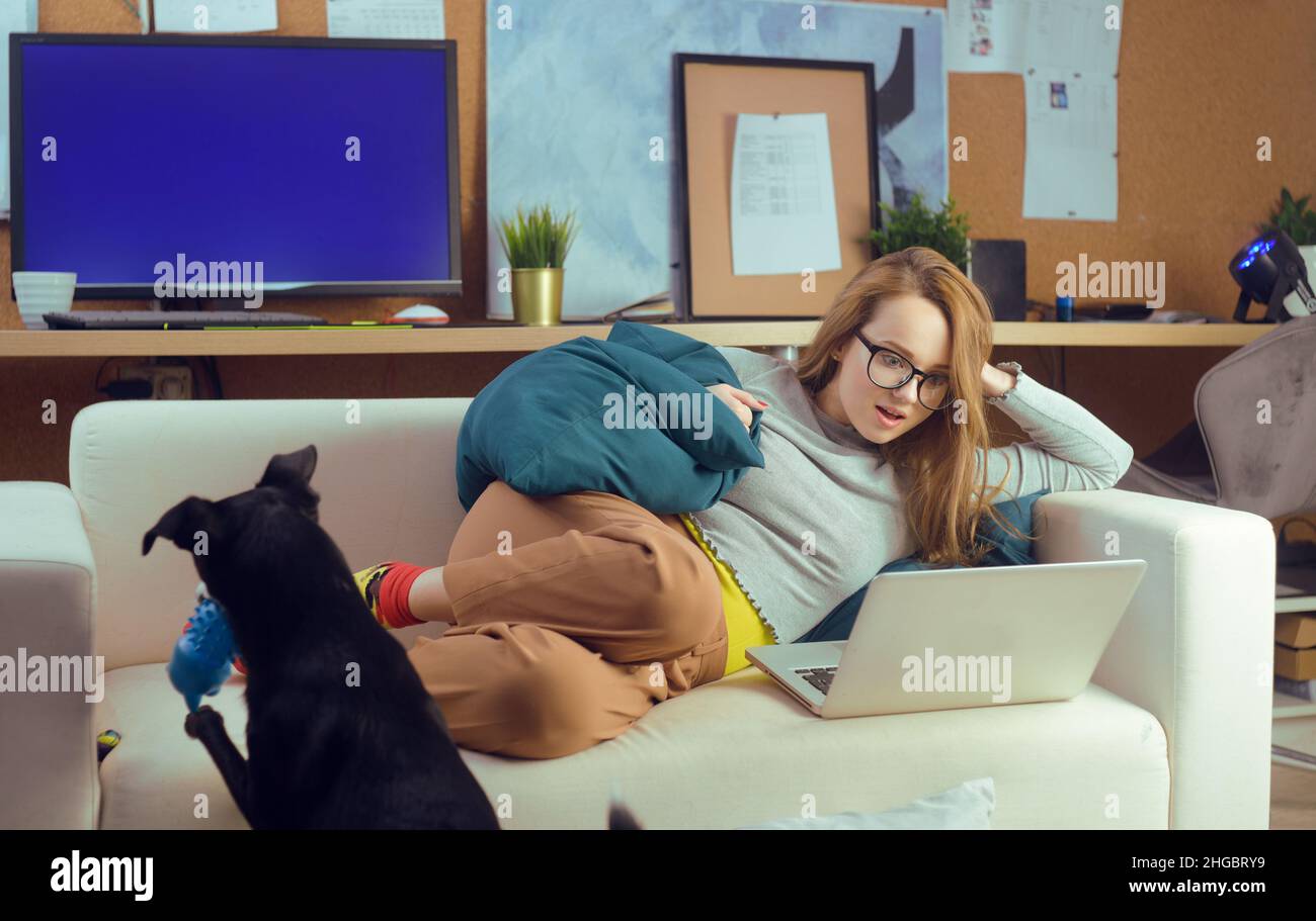 Ragazza sdraiata su un divano guardando un computer portatile. La ragazza si trova sul devan e guarda programmi TV, video, procrastinazione. Foto Stock