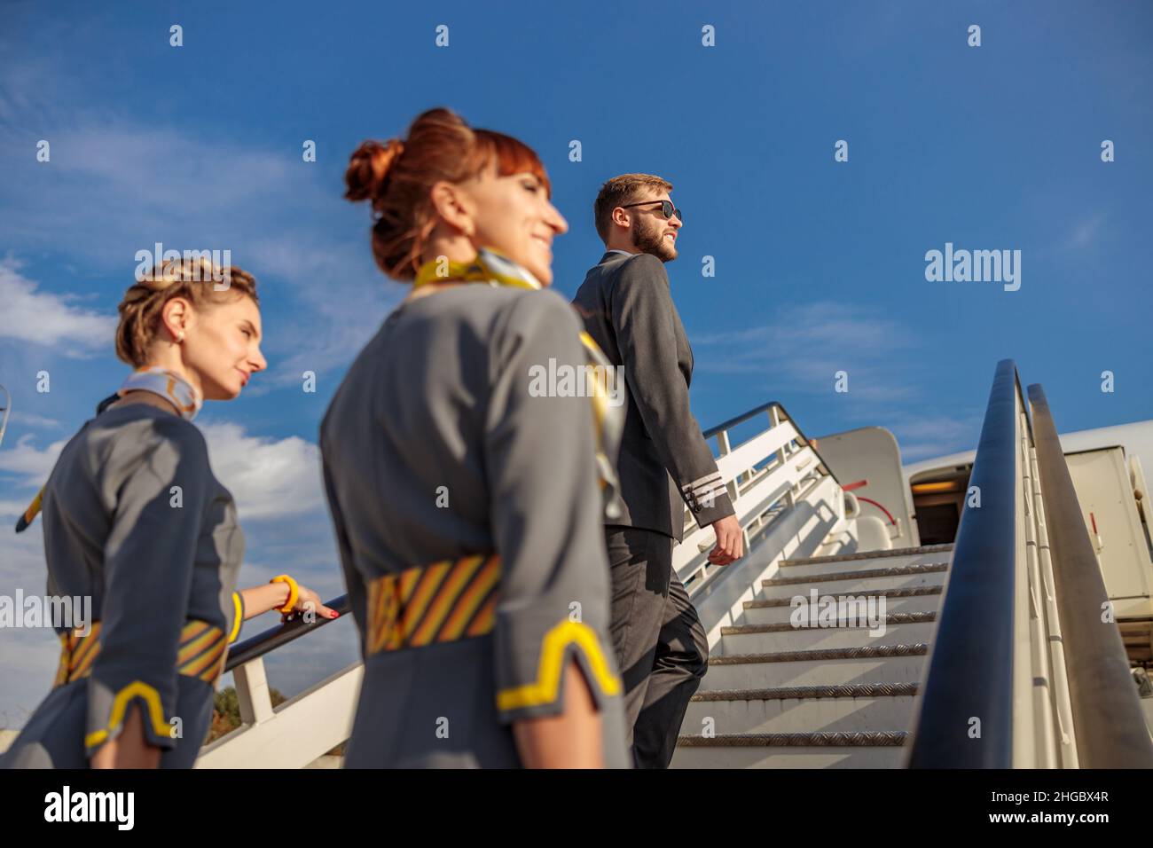 Uomo sorridente capitano dell'aviazione e due donne stewardesses salire piano di imbarco scale sotto il cielo blu Foto Stock