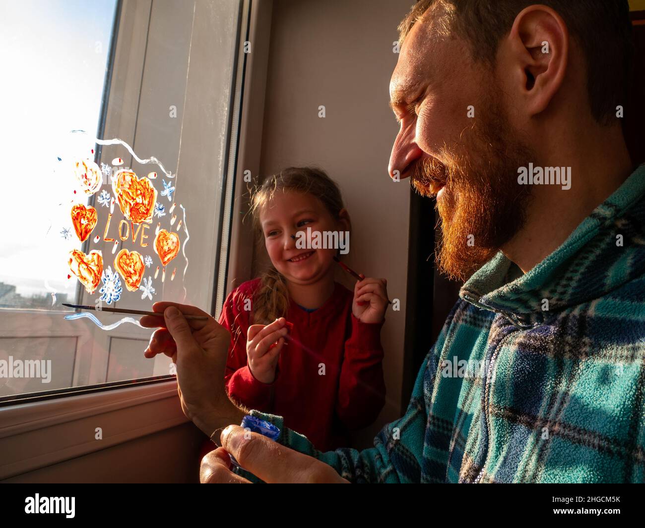 Bambino con padre pittura segno d'amore, giocando al chiuso quarantena famiglia tempo libero. La bambina tiene il pennello in mano e richiama il cuore rosso sul vetro della finestra. Foto Stock