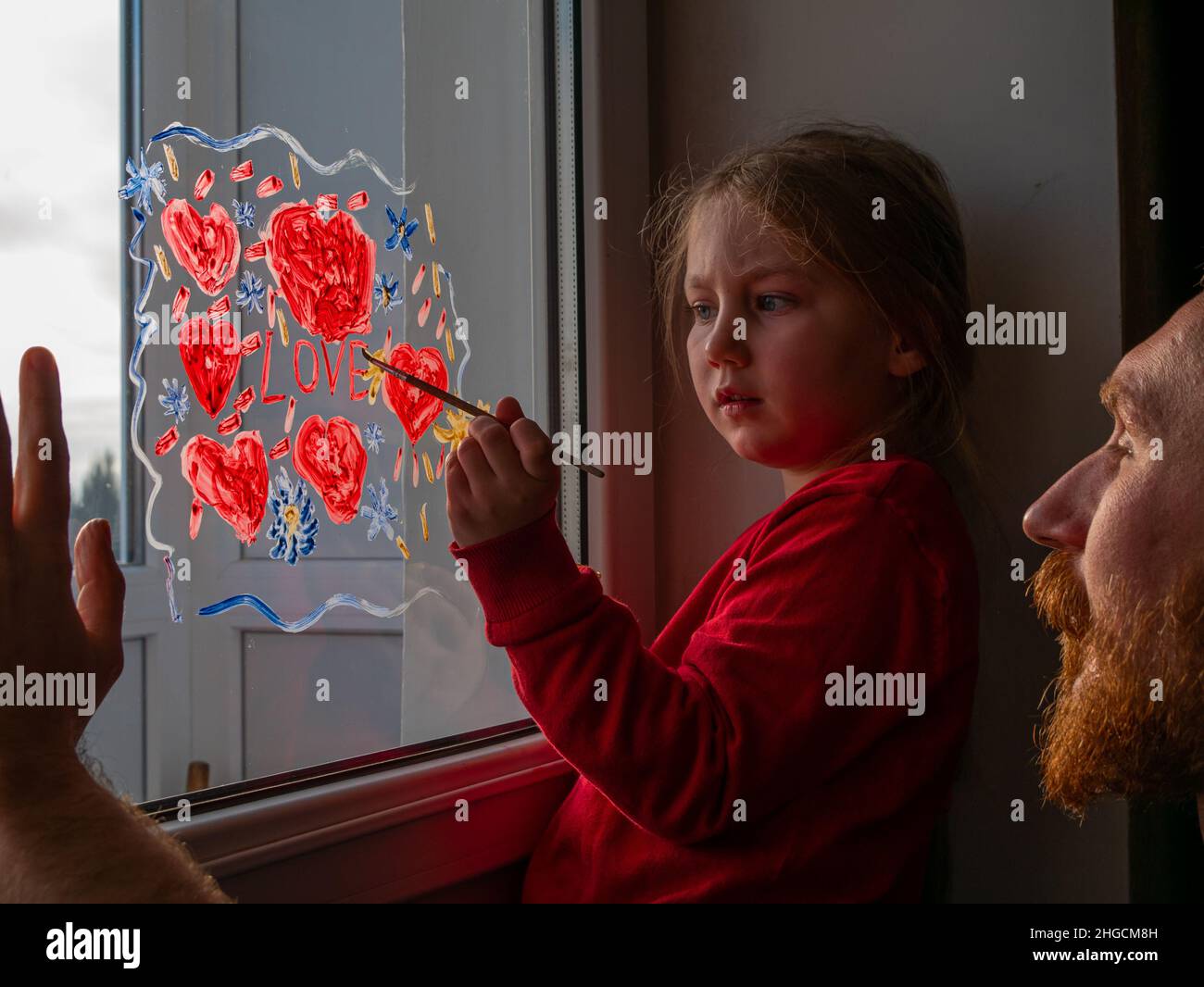 Bambino con padre pittura segno d'amore, giocando al chiuso quarantena famiglia tempo libero. La bambina tiene il pennello in mano e richiama il cuore rosso sul vetro della finestra. Foto Stock