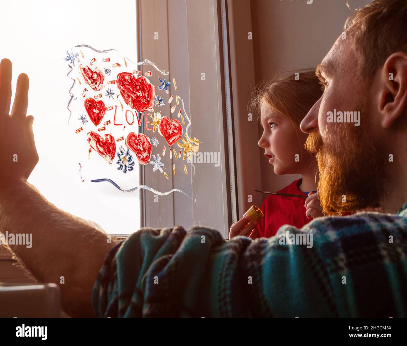 Bambino con padre pittura segno d'amore, giocando al chiuso quarantena famiglia tempo libero. La bambina tiene il pennello in mano e richiama il cuore rosso sul vetro della finestra. Foto Stock