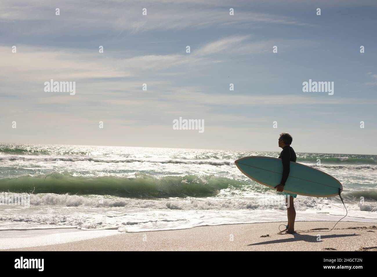 L'intera lunghezza di uomo anziano biraciale che trasporta la tavola da surf guardando le onde di spruzzi in mare in giorno di sole Foto Stock