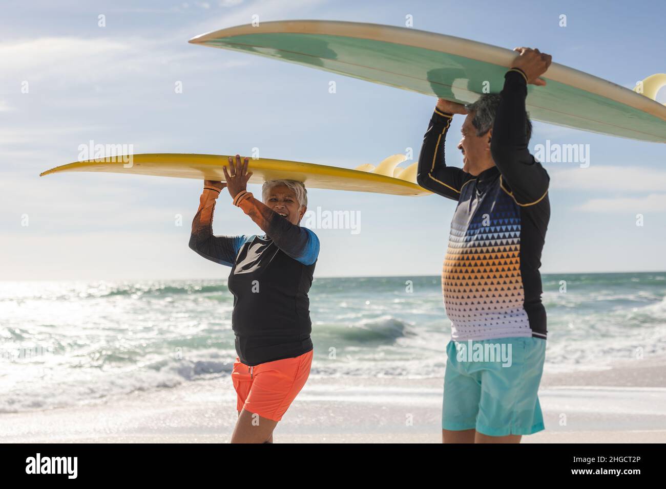 Coppia anziana multirazziale felice che si guardano mentre trasportano tavole da surf sulla testa alla spiaggia Foto Stock