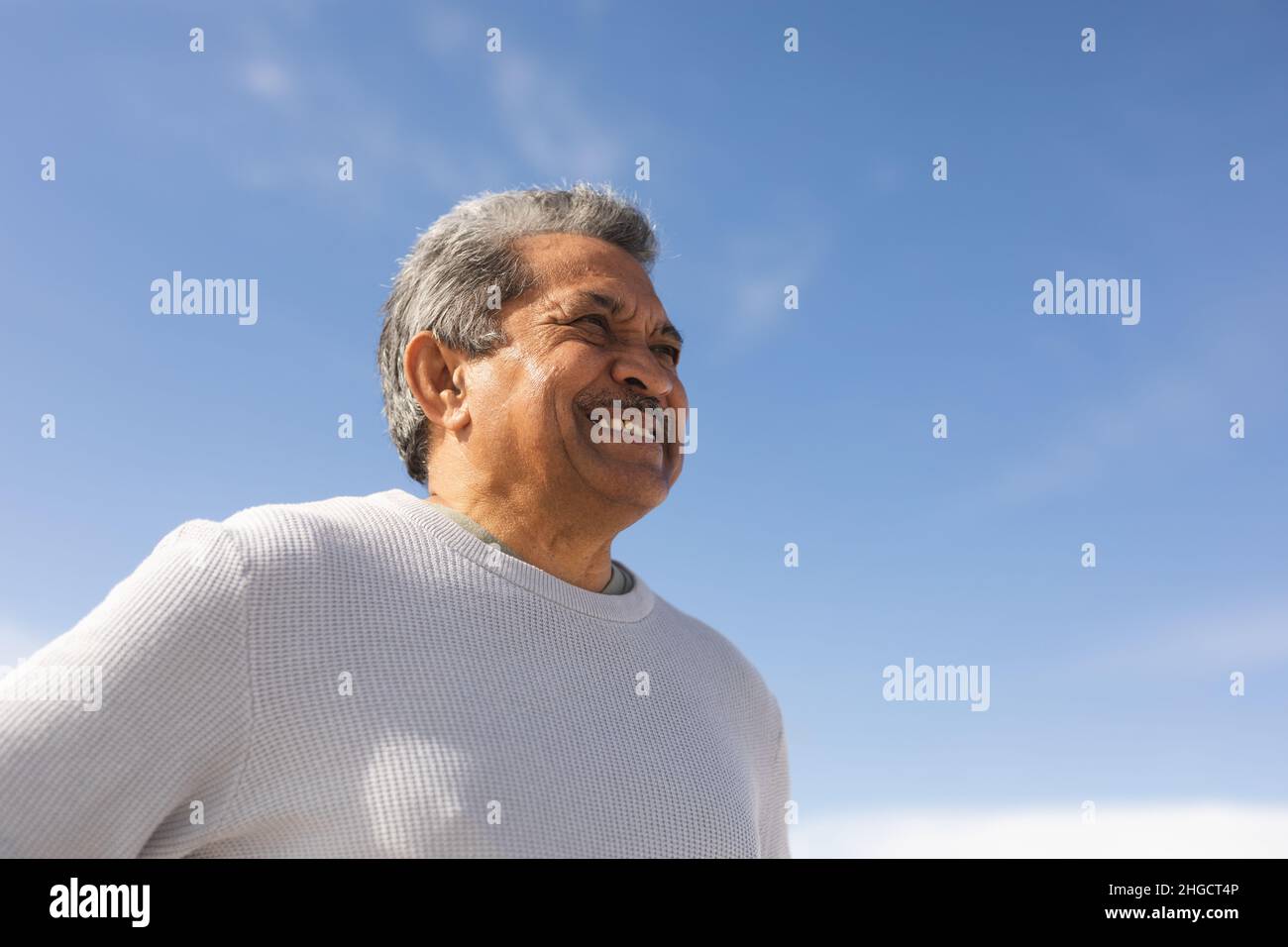 Vista ad angolo basso dell'uomo anziano biraciale sorridente che guarda via alla spiaggia contro il cielo blu nelle giornate di sole Foto Stock