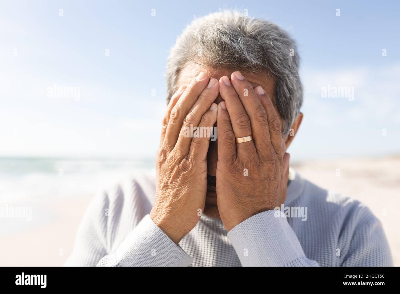 Primo piano di un uomo anziano biraciale preoccupato che copre il viso con le mani in spiaggia contro il cielo nelle giornate di sole Foto Stock