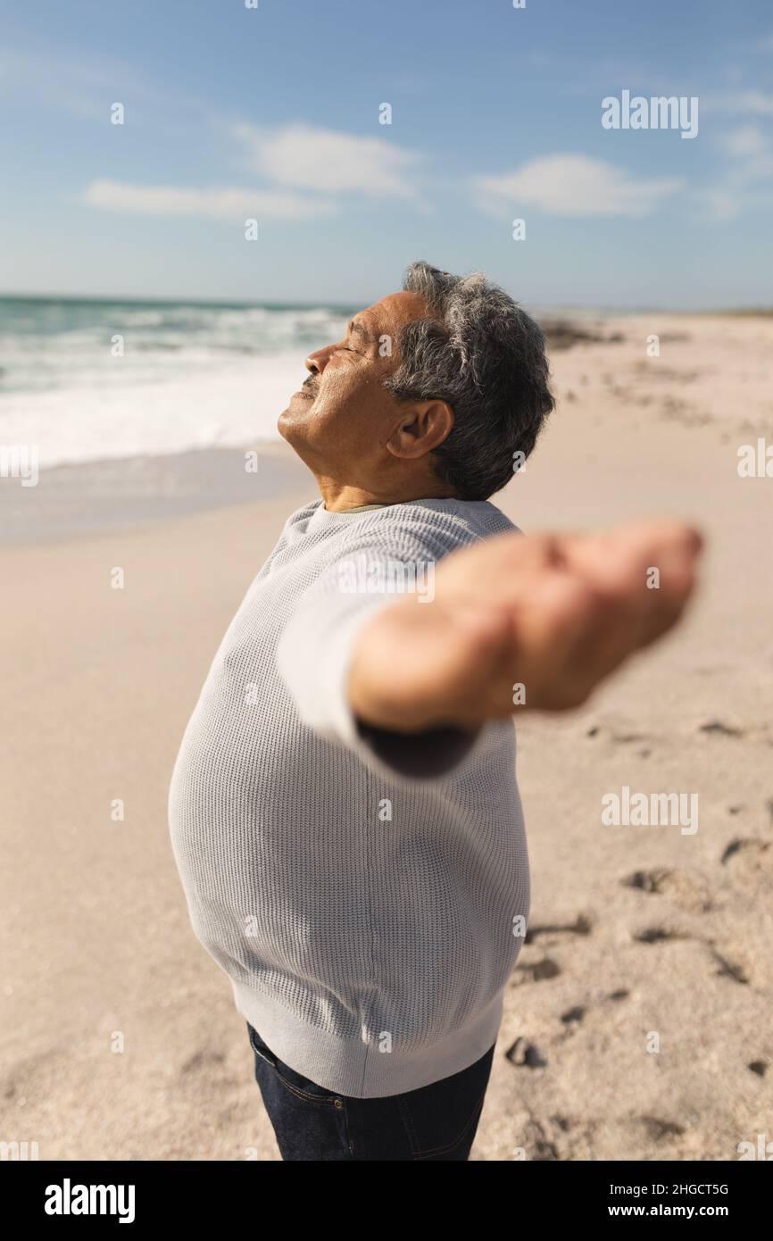Vista laterale dell'anziano biraciale con gli occhi chiusi e le braccia distese in spiaggia il giorno di sole Foto Stock