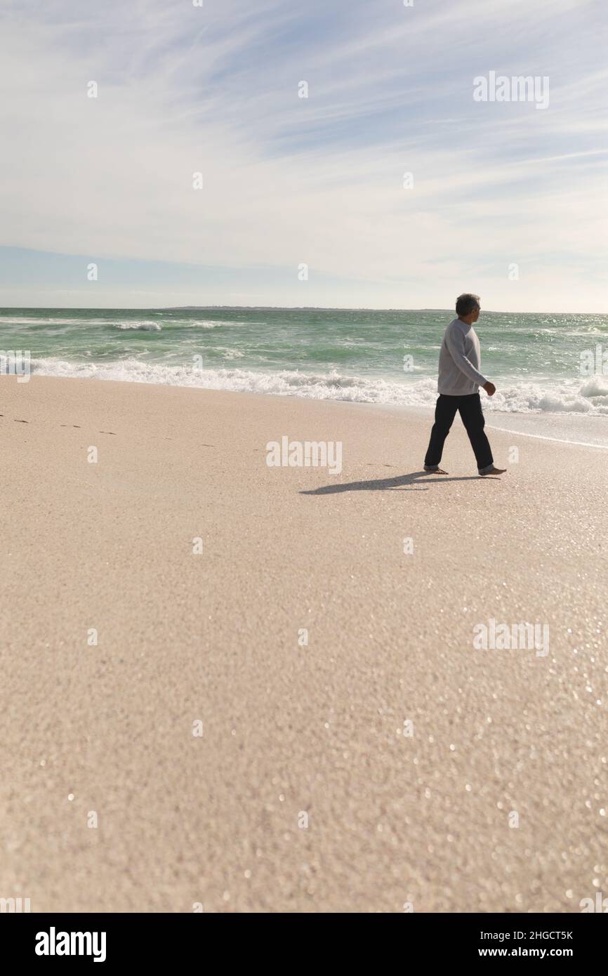 L'intera lunghezza dell'anziano biraciale in pensione che cammina sulla spiaggia contro il cielo durante la giornata di sole Foto Stock