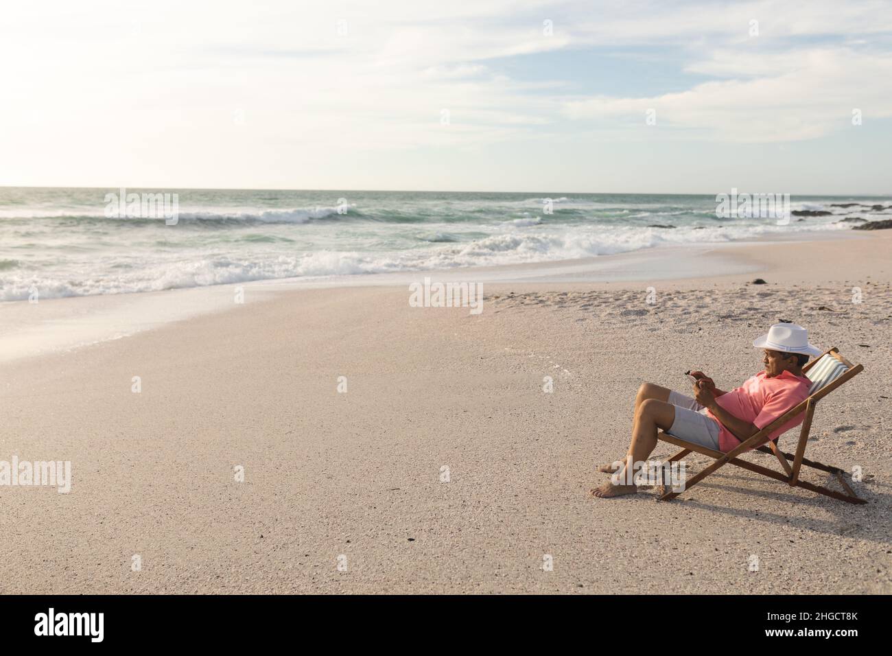 Vista laterale completa dell'uomo anziano biraciale utilizzando un tablet digitale rilassante su una sedia pieghevole in spiaggia Foto Stock