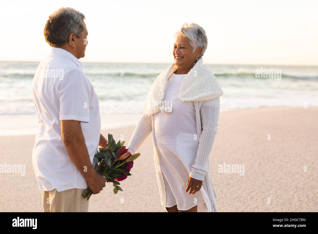 Sorridente donna biraciale anziana che tiene la mano dell'uomo con il bouquet alla spiaggia durante la cerimonia nuziale Foto Stock