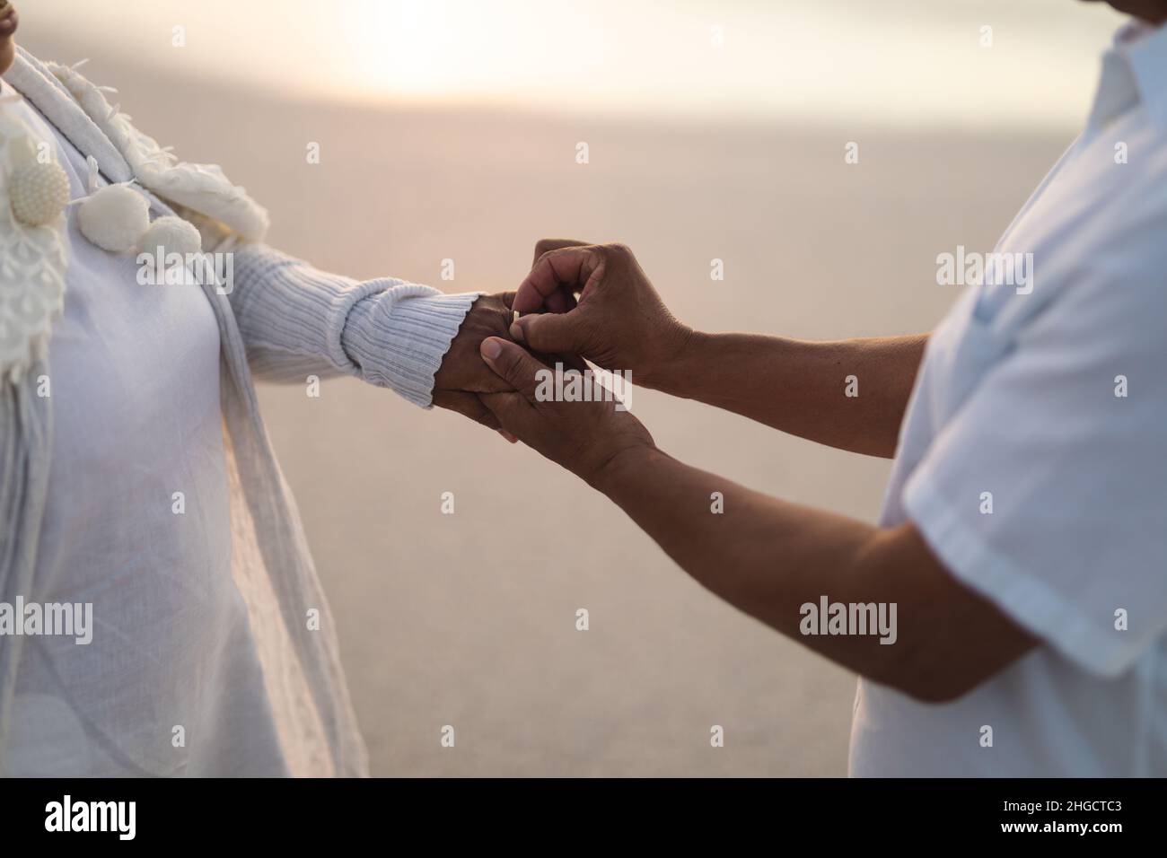 Sezione centrale dello sposo superiore del biracial che mette l'anello di nozze sul dito della sposa alla spiaggia durante il tramonto Foto Stock