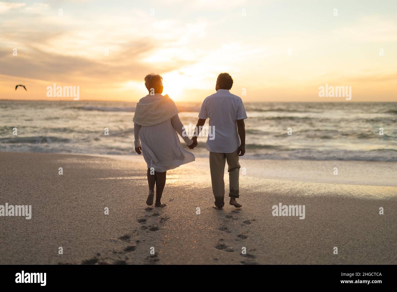 Tutta la lunghezza della coppia multirazziale newlywed anziana che cammina verso la riva alla spiaggia durante il tramonto Foto Stock