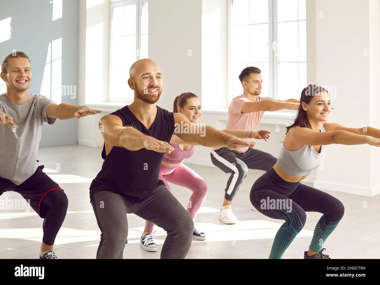 Gruppo di giovani felici che si allenano insieme in posizione squat in una palestra spaziosa e luminosa. Foto Stock Gruppo di giovani felici che si allenano insieme in posizione squat in una palestra spaziosa e luminosa. Foto Stock