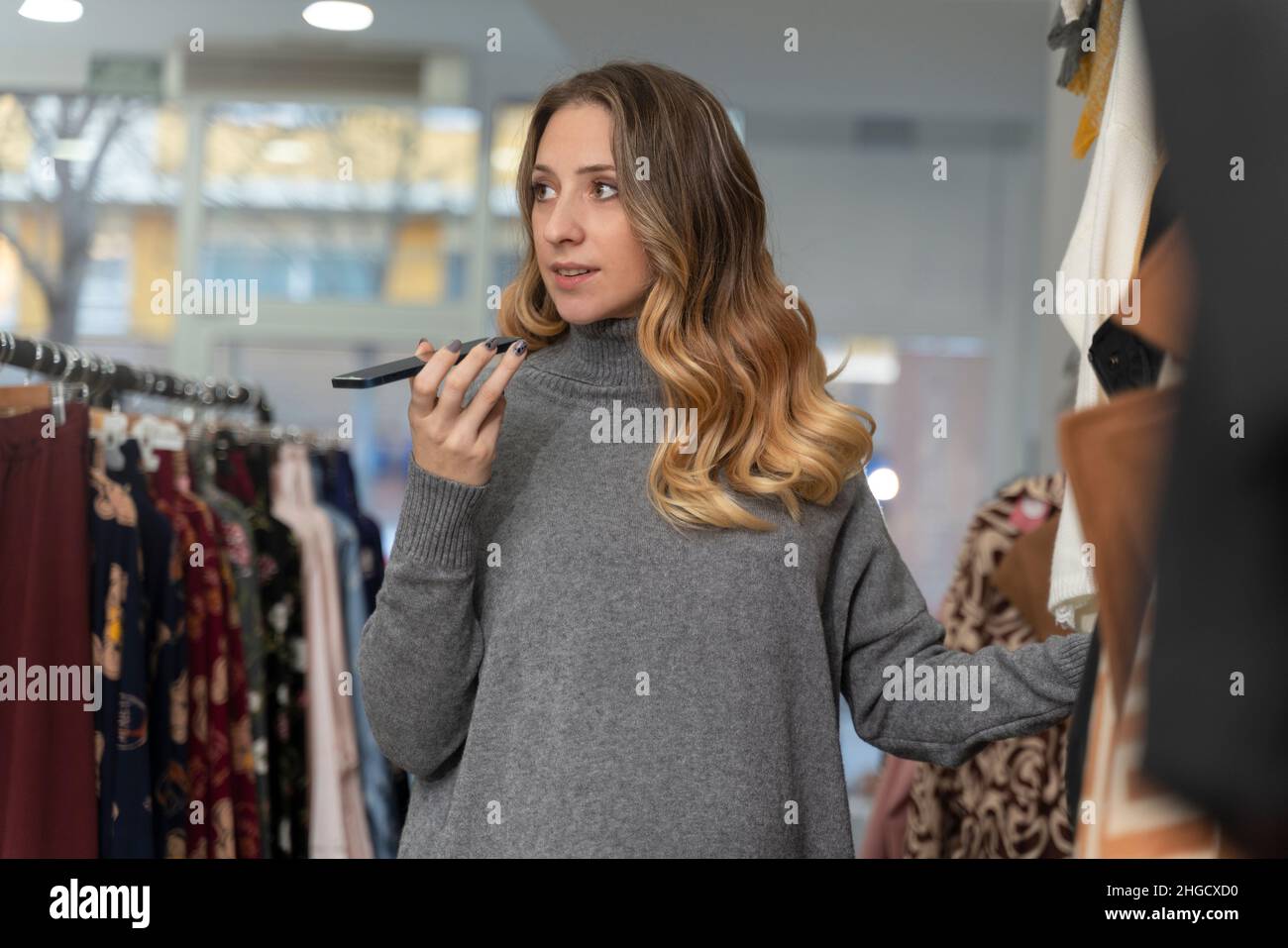 Giovane donna bionda in cerca di vestiti in un negozio e inviare un messaggio audio Foto Stock