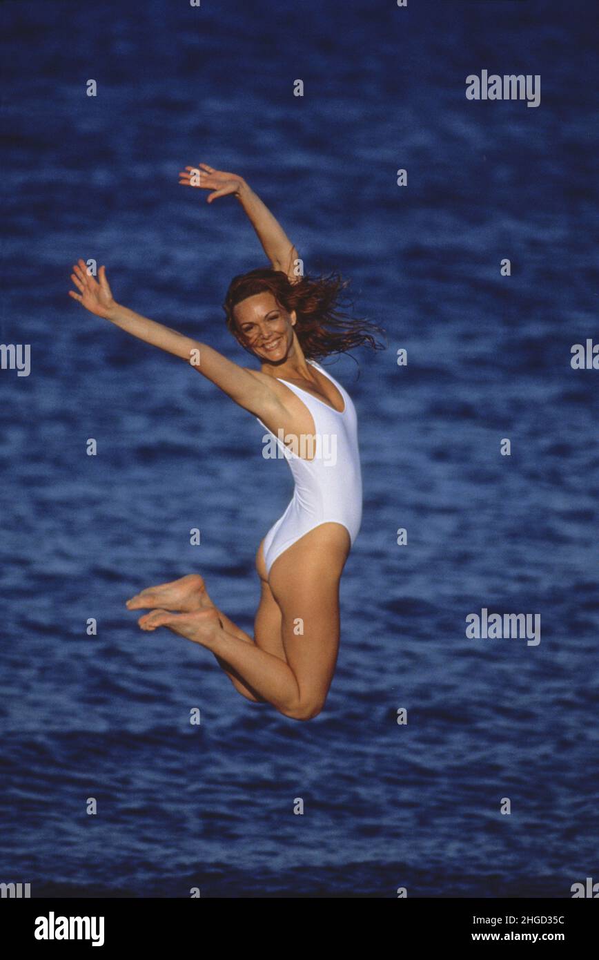 Atletica giovane donna felice sorridente facein bianco tuta bathin jumping sulla sabbia blu mare sulla terra posteriore Foto Stock