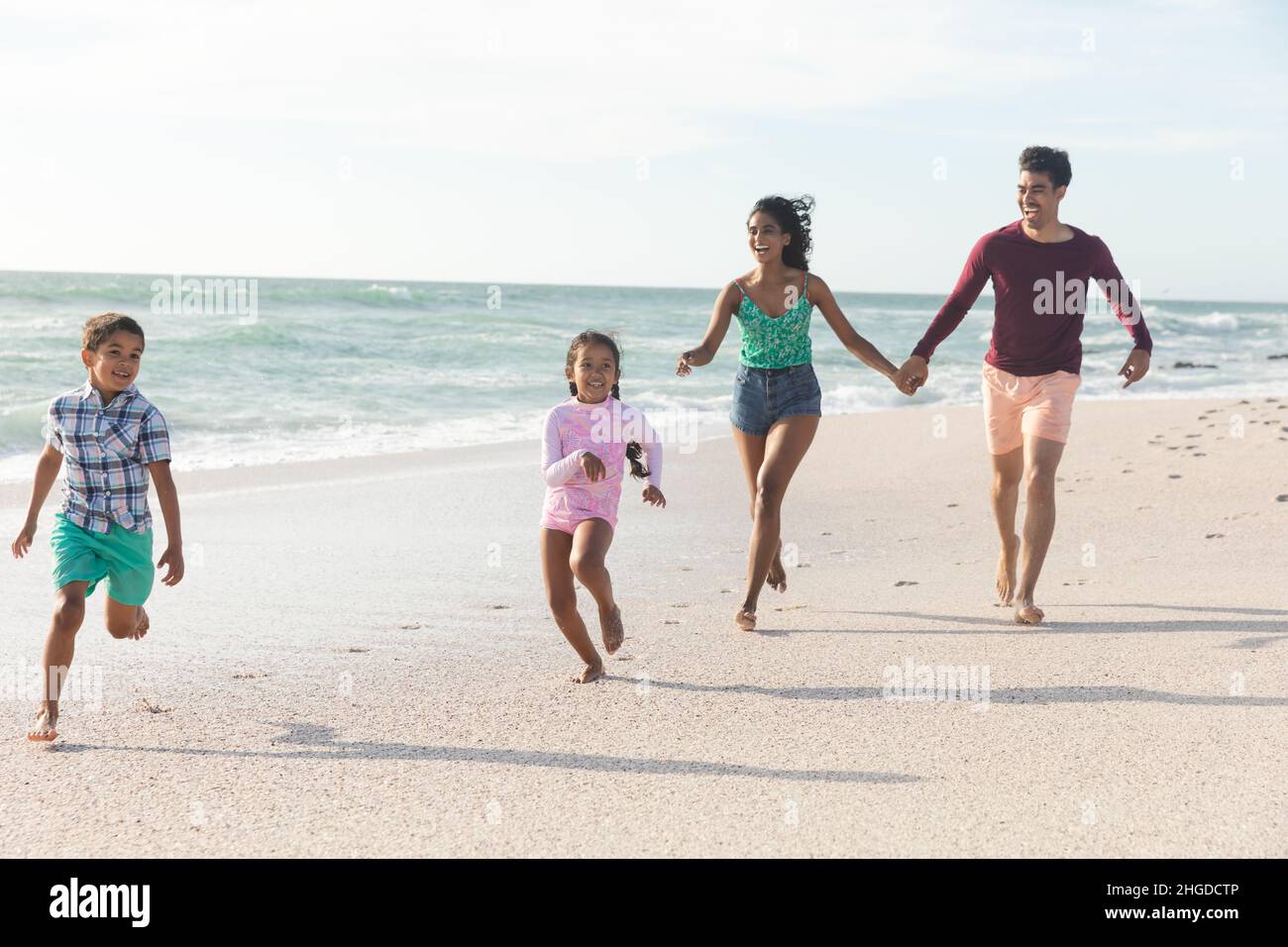 Tutta la lunghezza della famiglia multirazziale che corre insieme sulla spiaggia contro il cielo durante la giornata di sole Foto Stock