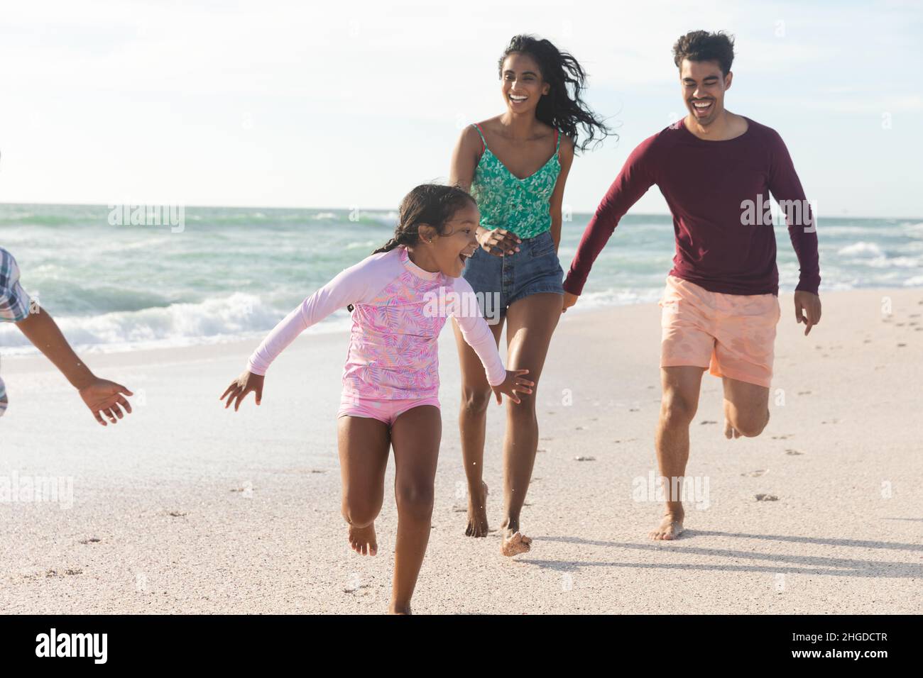 Allegra famiglia multirazziale che corre sulla spiaggia contro il cielo durante la giornata di sole Foto Stock