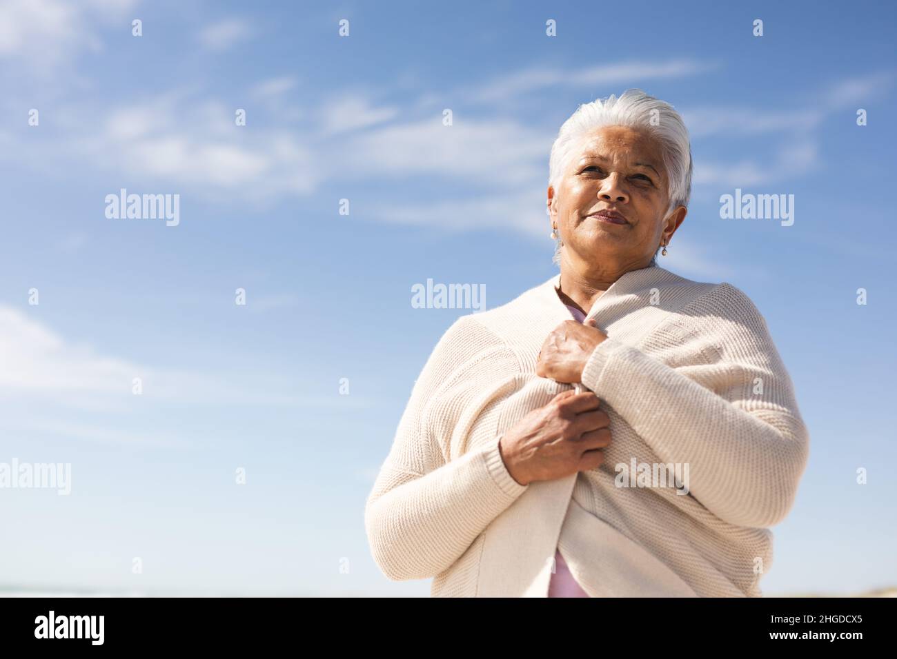 Vista ad angolo basso della donna anziana biraciale che indossa un tappeto guardando via alla spiaggia contro il cielo blu Foto Stock