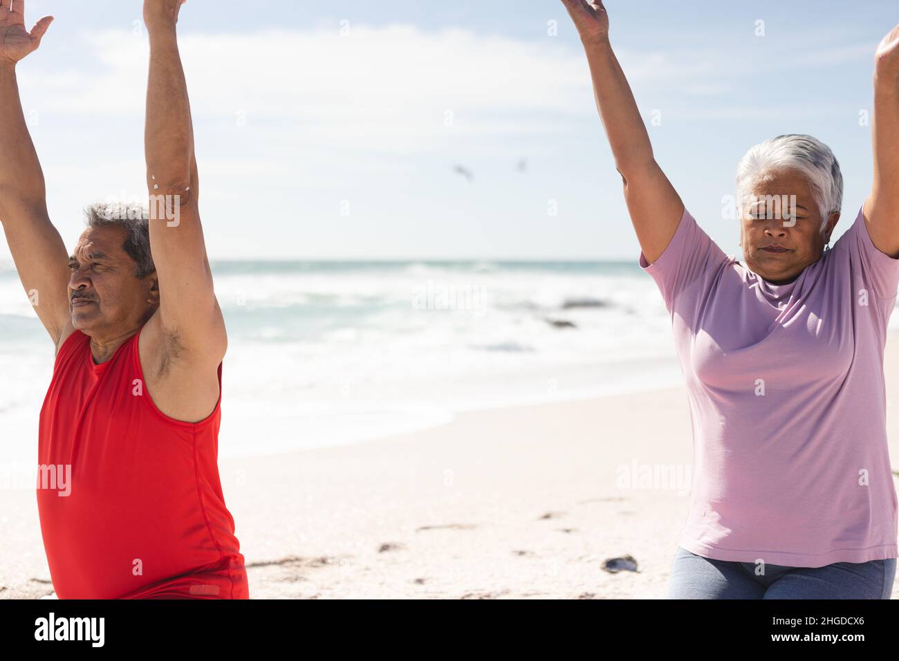 Coppia biraciale senior che si esercita insieme con le braccia sollevate in spiaggia contro il cielo in giorno di sole Foto Stock