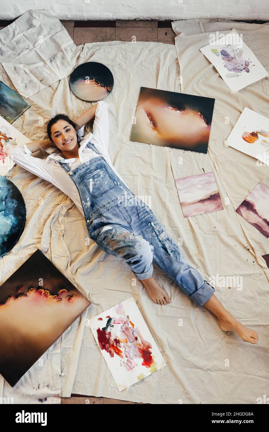 Vista dall'alto di un'artista femminile sdraiata al centro dei suoi dipinti. Felice giovane pittore sorridente alla macchina fotografica nella sua galleria d'arte. Creatività giovane WO Foto Stock