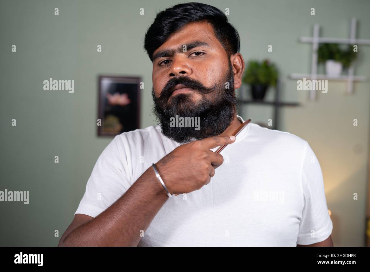 Il giovane uomo pettina la barba guardando la macchina fotografica a casa - concetto di cura del corpo, cura del corpo e benessere Foto Stock