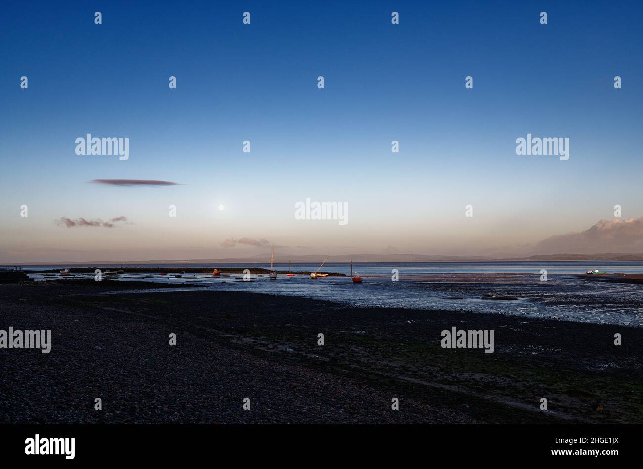 La luna nel cielo del mattino sopra le barche sul fango in Morecambe Bay, Lancashire, quando la marea è fuori, mattina presto. Foto Stock