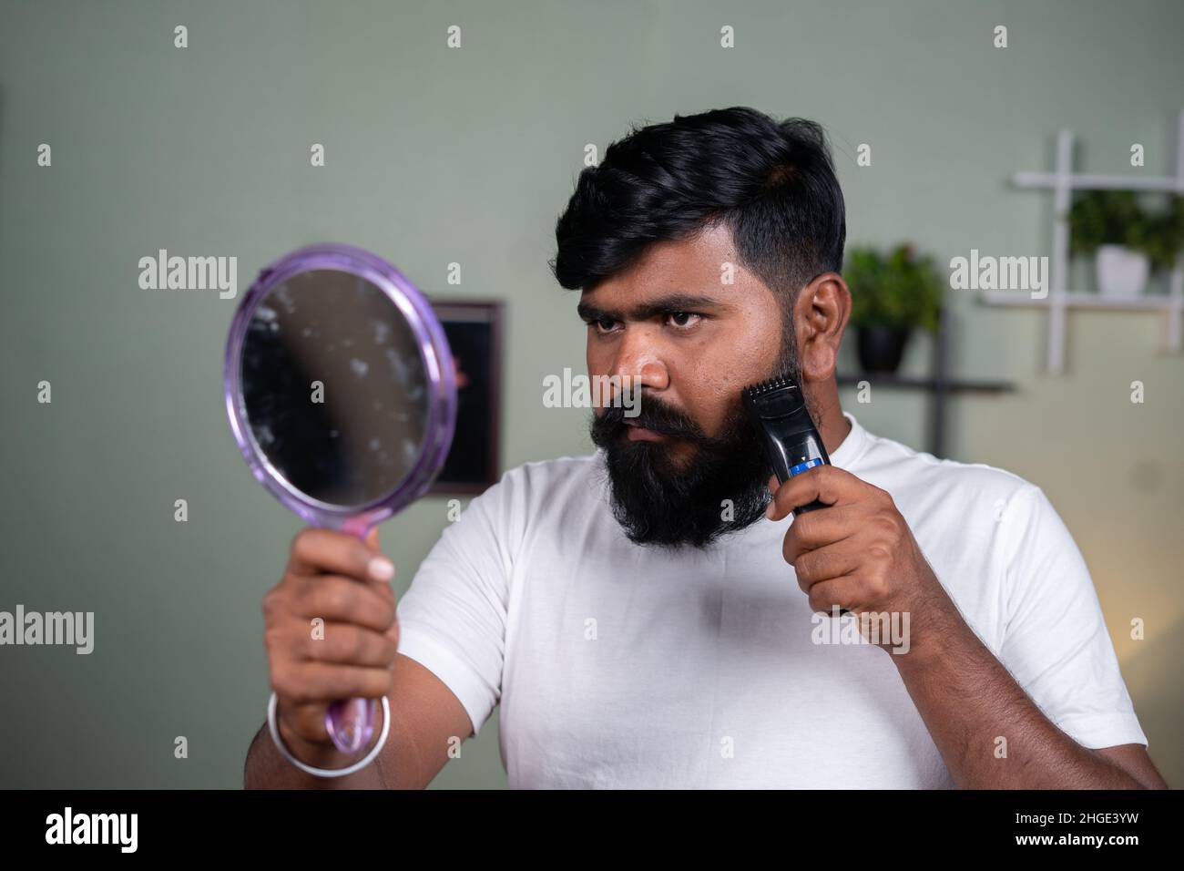 Giovane uomo indiano della barba che usa il rasoio elettrico o la macchina di trimbere guardando il concetto specchio- della cura dei capelli, del grooming e del wellness. Foto Stock