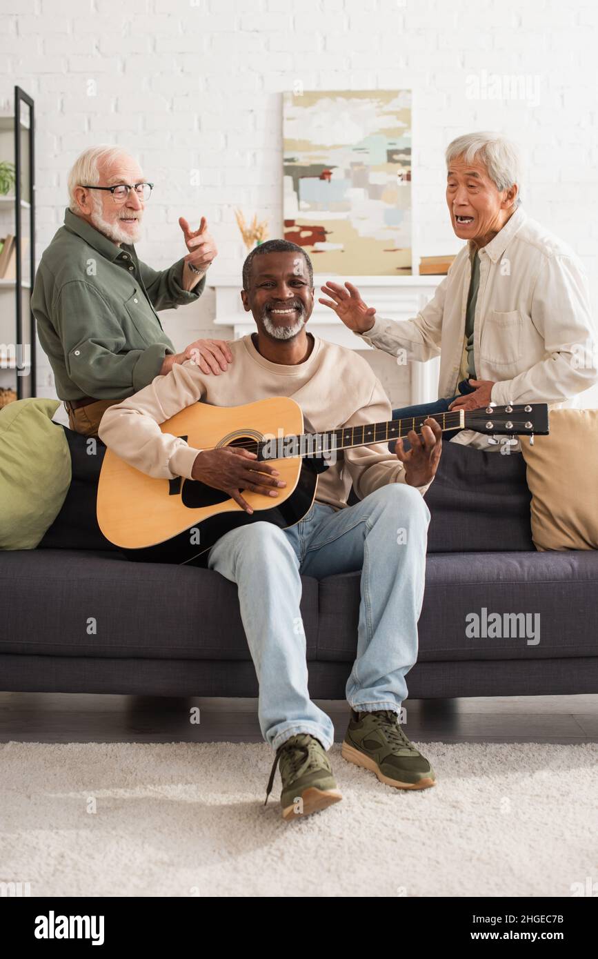 Uomini senior interrazziali cantano vicino a un amico afroamericano che suona la chitarra acustica in casa Foto Stock