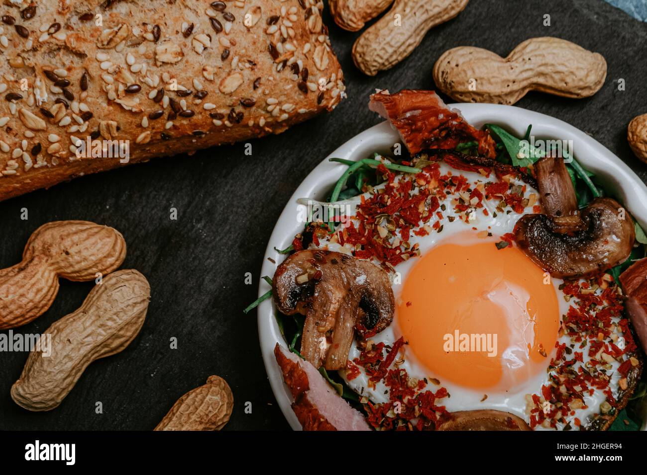 Uova fritte cucinate perfettamente con funghi, spezie, pepe, carne e sale su rucola. Piatto colazione rotondo su tavola nera con arachidi e baguette w Foto Stock