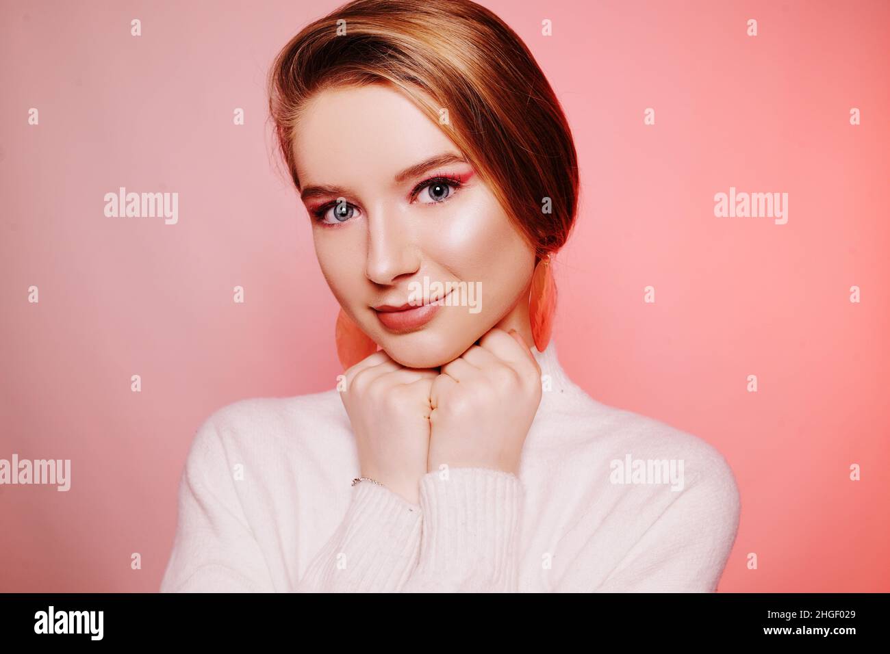 Biondo incredibilmente bello su sfondo rosa. Ritratto del modello del viso del bambino. Ragazza con capelli leccati e grandi orecchini. Gioielli. Ragazza in un sudore bianco Foto Stock