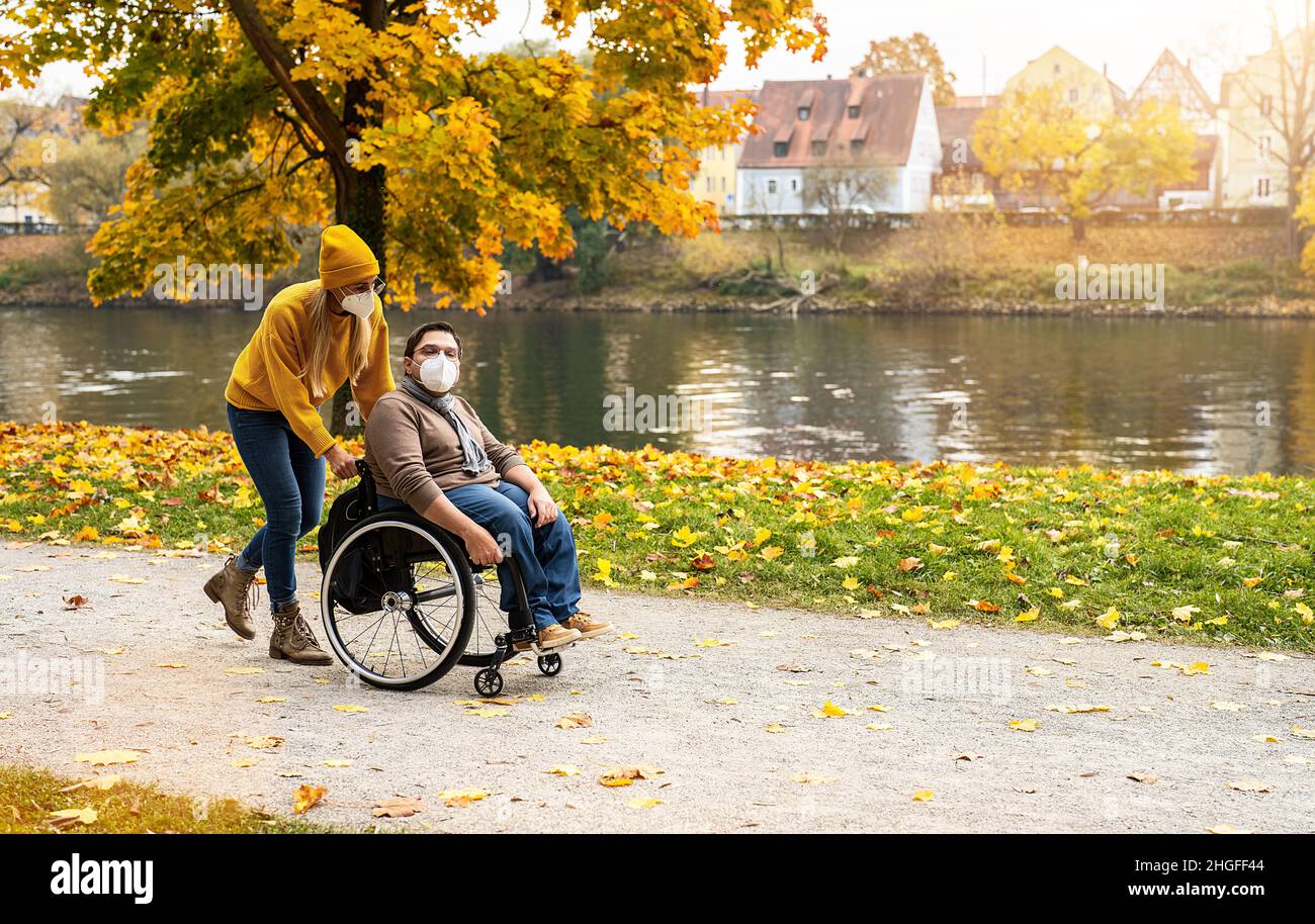 Donna e la sua amica con maschera facciale in sedia a rotelle che hanno passeggiare nel parco in un fiume godendo l'autunno durante la pandemia del convivio-19 Foto Stock