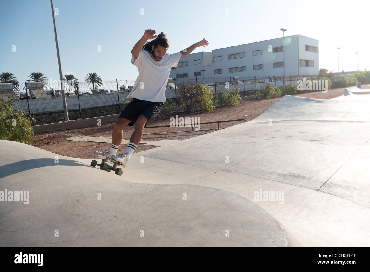 Skater maschile giovane adulto che cavalca sulle rampe del parco skate. Indossa abiti estivi e ha lunghi capelli neri. Foto Stock
