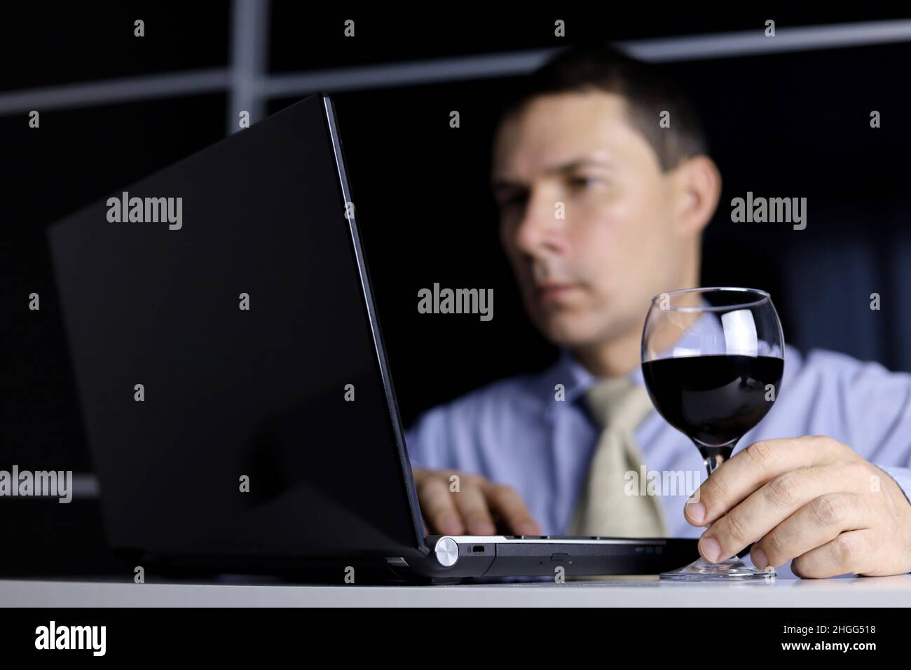 Uomo con un bicchiere di vino rosso in mano, seduto al computer portatile. Lavorate a distanza a casa o rilassatevi in ufficio con una bevanda alcolica Foto Stock