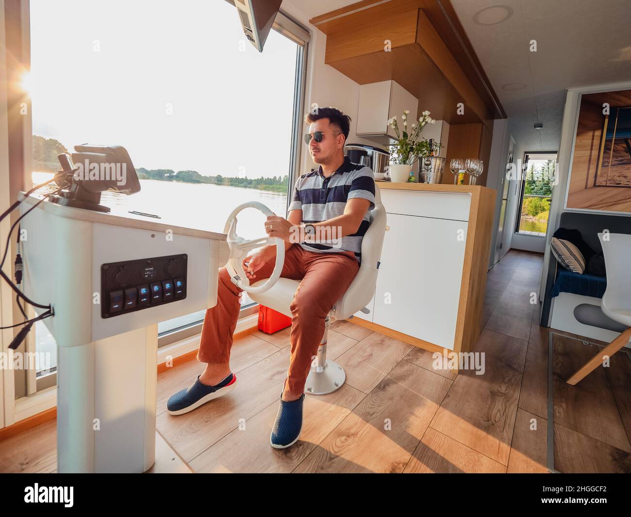 Un uomo che indossa abbigliamento casual con capelli neri che guidano casa galleggiante completamente attrezzata su un fiume durante le sue vacanze estive in una giornata di sole. Viaggiare sul bar Foto Stock