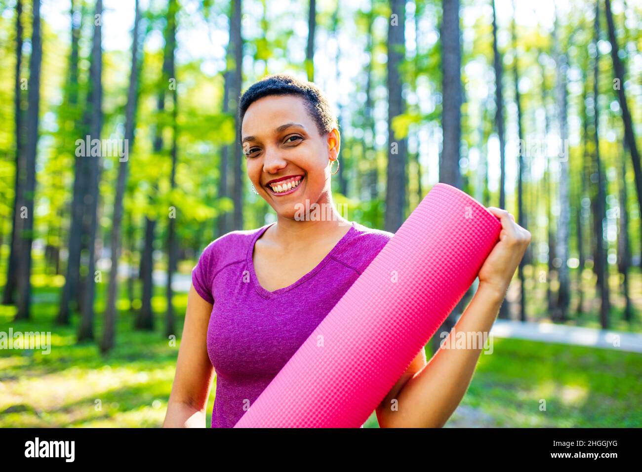 Giovane allenatore afro-americano felice con capelli corti tenendo un tappeto yoga all'aperto e guardando la macchina fotografica Foto Stock